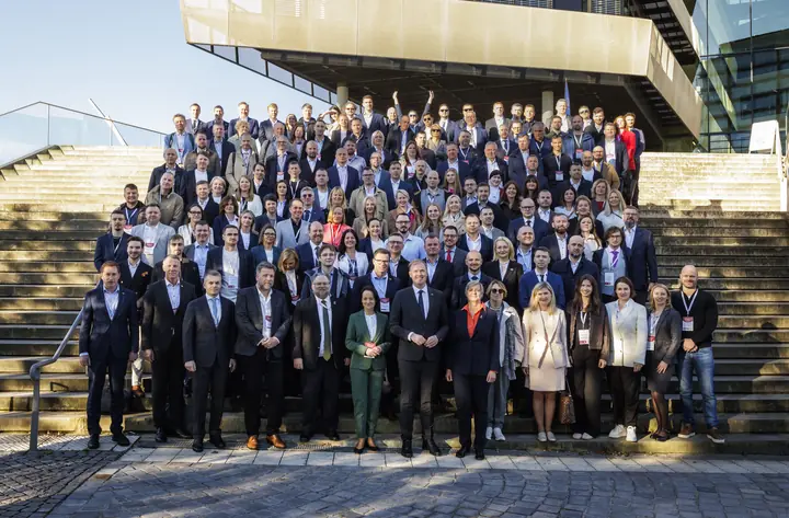 The full Latvian delegation of more than 180 people on the wide steps in front of a modern building with a cantilevered upper floor, in low autumn sun.