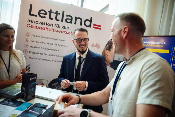 Aleksandrs Gusevs of Double Helix Technologies handing a card to a visitor at the Latvian stand, under the banner "Lettland, Innovation für die Gesundheitswirtschaft".