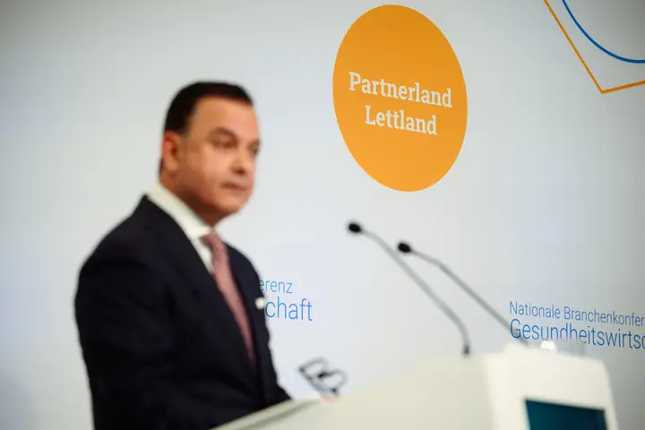 The orange "Partnerland Lettland" badge on the conference backdrop, with a speaker at the lectern out of focus in the foreground.