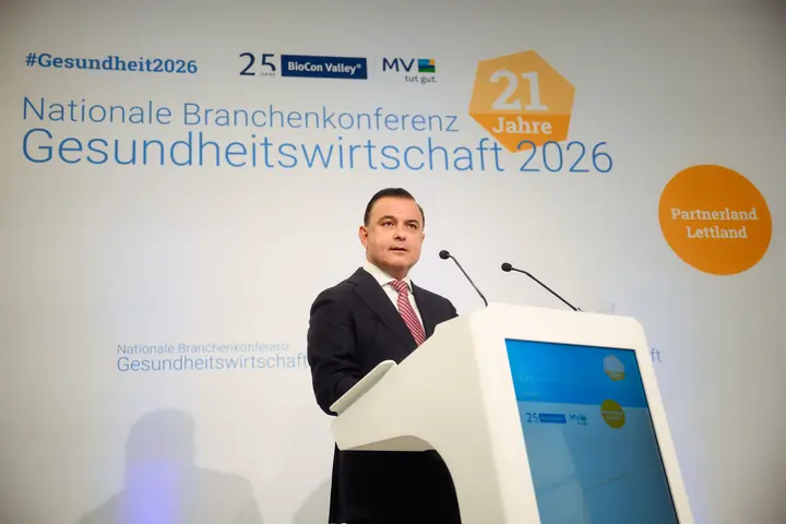 A speaker at the conference lectern in front of the backdrop reading "Nationale Branchenkonferenz Gesundheitswirtschaft 2026" and "Partnerland Lettland".