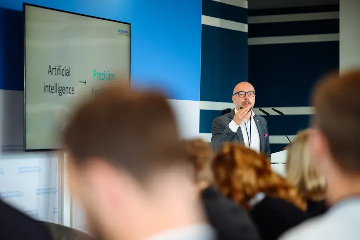 Armands Baranovskis of Double Helix Technologies presenting to the audience; the screen reads "Artificial intelligence" with an arrow to "Precision health".