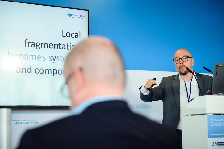 Armands Baranovskis, CEO of Double Helix Technologies, at the lectern; the screen beside him reads "Local fragmentation becomes systemic and compounds" under the Double Helix Technologies logo.