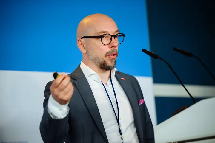 Armands Baranovskis, CEO of Double Helix Technologies, speaking at the conference lectern with a presenter remote in his hand.