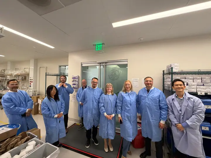 Delegation members in blue laboratory coats and safety glasses during the tour of the Thermo Fisher Scientific site.